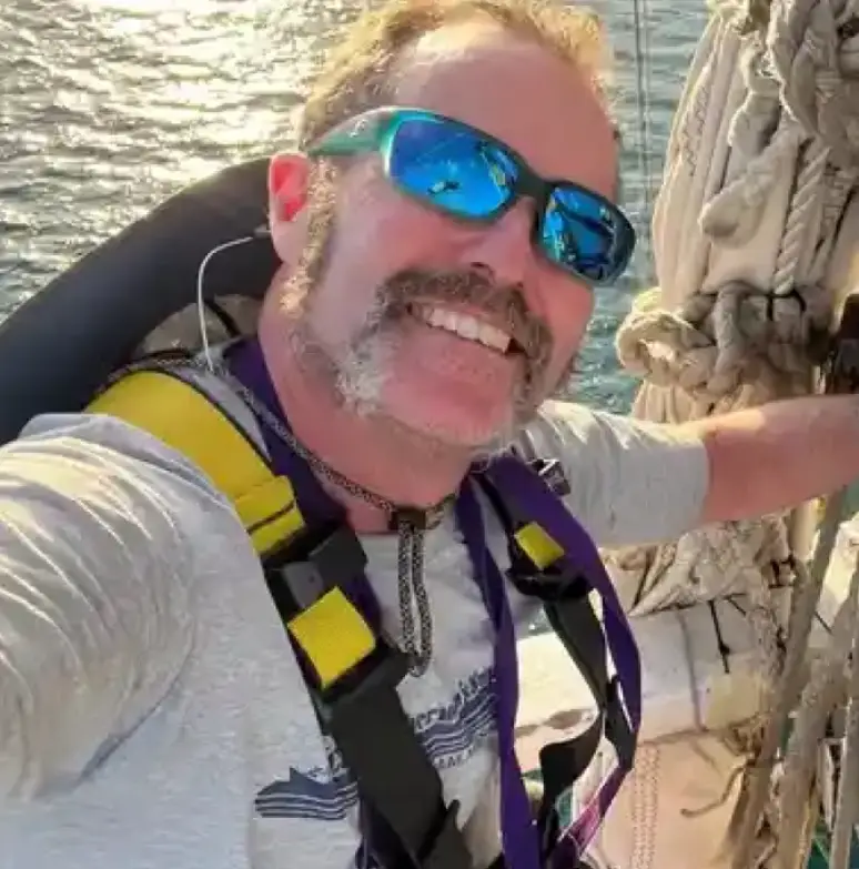 Man wearing a t-shirt and blue sunglasses, while taking a selfie on a sailboat in the water