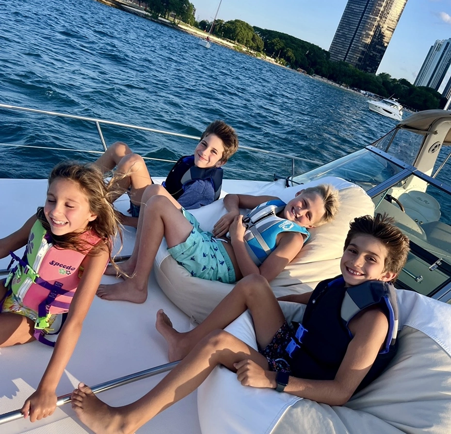 Group of four children sitting on top of a boat in the water, surrounded by trees, buildings