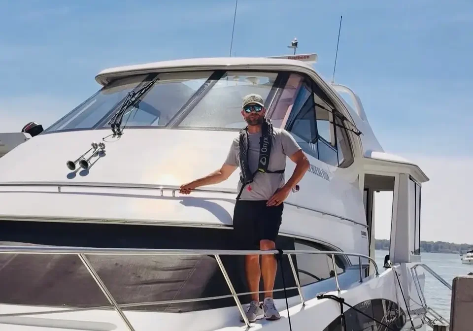 Man standing on the deck of a white boat, wearing a t-shirt, black shorts, and a cap