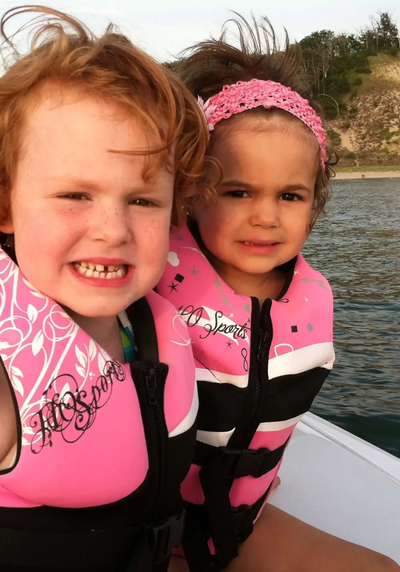 Two girls in life jackets sitting on a boat, and enjoying the view of the water, sand, trees, and sky