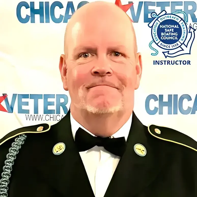 Man in a tuxedo and bow tie standing in front of a banner that reads "Veteran of the "